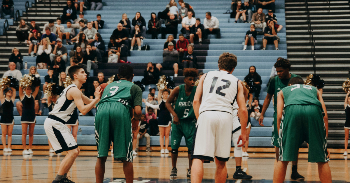 A middle school gym hosting a basketball game. Players on the court waiting for the play to start. Cheerleaders and fans in the stands. 